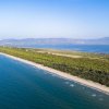 Lunga spiaggia sabbiosa con mare azzurro e vegetazione verde, monti in lontananza.