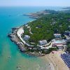 Una costa con spiaggia sabbiosa, mare azzurro e case tra alberi verdi.
