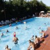 Piscina affollata con persone che nuotano e si rilassano su lettini, circondata da alberi.