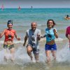 Cinque persone sorridenti corrono in mare, con il cielo azzurro sopra e vele sullo sfondo.