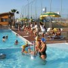Persone in piscina con docce, alcune sedute su sdraio sotto ombrelloni.