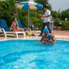 Piscina con persone che nuotano e una donna su una sdraio sotto un ombrellone.