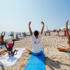 Persone fanno yoga sulla spiaggia, sedute su asciugamani con le braccia alzate.