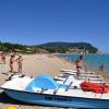 Spiaggia con pedalò, persone in costume e colline verdi sullo sfondo.