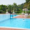 Piscina all'aperto con persone che nuotano, circondata da sedie bianche e palme sullo sfondo.