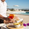 Una persona in camicia bianca tiene un drink arancione vicino a una piscina con snack e fiori.