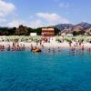 Spiaggia affollata con bagnanti e sdraio verdi, mare blu in primo piano, montagne sullo sfondo.