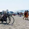 Biciclette parcheggiate sulla spiaggia, persone e ombrelloni sullo sfondo.