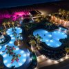 Piscine illuminate di notte con palme e giochi d'acqua, spiaggia sullo sfondo.