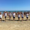 Persone in fila sulla spiaggia partecipano ad attività, con il mare sullo sfondo.