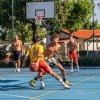 Giovani giocano a basket su un campo all'aperto, alcuni a torso nudo, con un canestro sullo sfondo.
