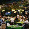 Persone sedute a tavoli verdi all'aperto in un ristorante, illuminato da luci e lanterne.