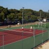Campo da tennis all'aperto con persone che giocano, circondato da alberi e recinzioni.