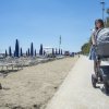 Donna con passeggino su una passeggiata vicino alla spiaggia, ombrelloni blu e sdraio sulla sabbia.