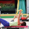 Persone sollevano pesi su tappetini da ginnastica, un istruttore guida davanti a un banner Zumba.